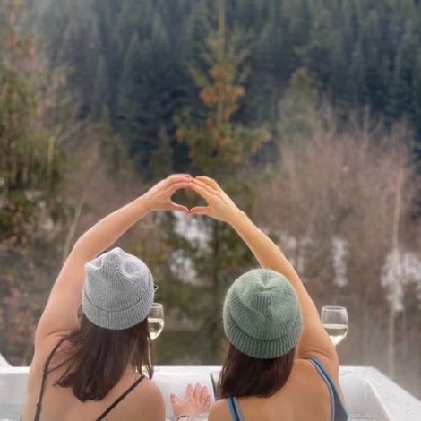 Dwie osoby w jacuzzi układają dłonie w kształt serca na tle zimowego lasu